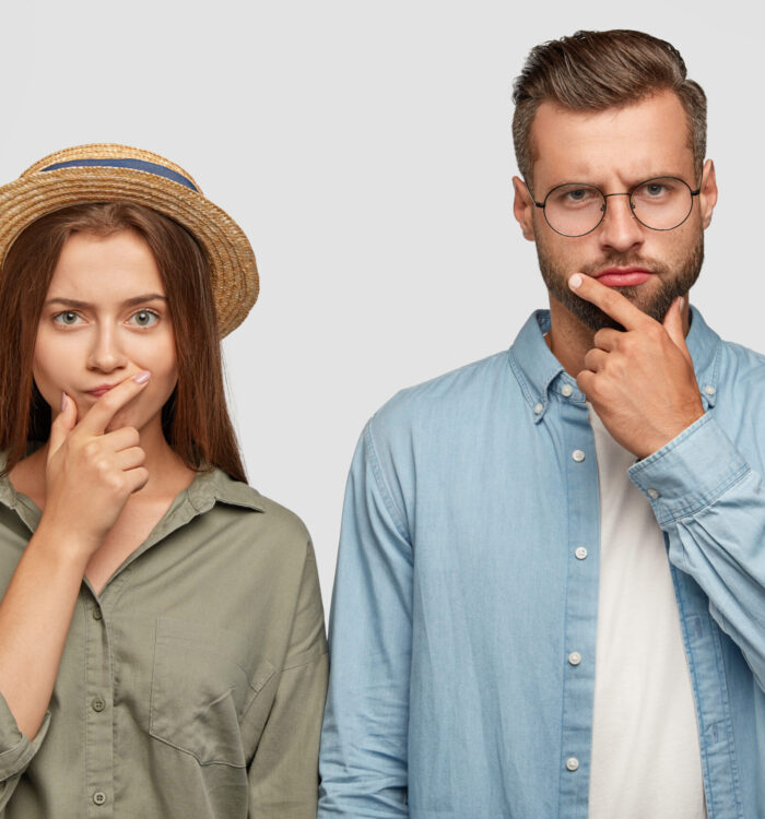 Photo of thoughtful confused female and male coworkers keep hands on chins, think how solve problem with debts, dressed in fashionable shirts, model in studio togeher. Hmm, let us think for moment